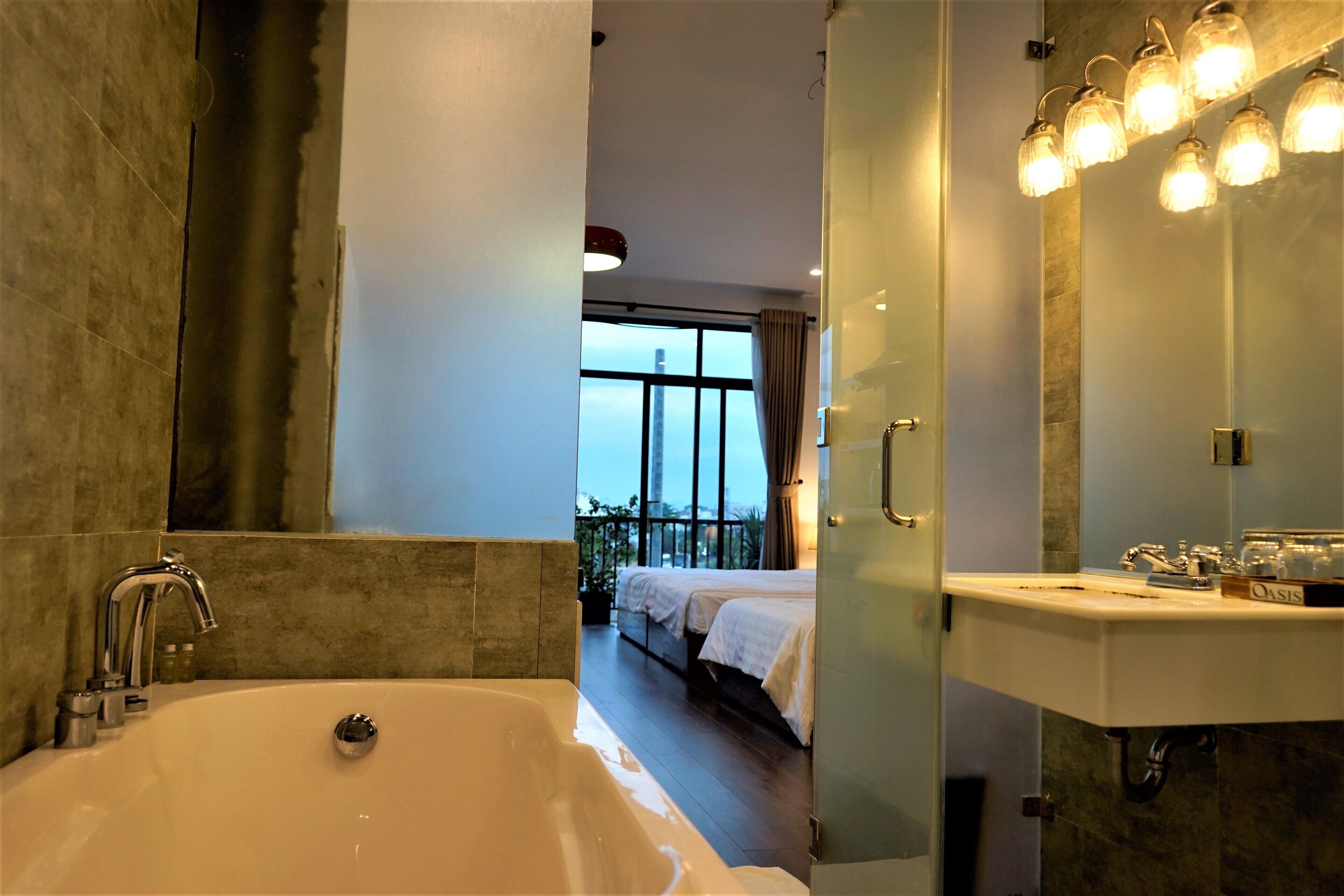 A modern bathroom with a bathtub, a large mirror with lights, and a view of a balcony with a bed.