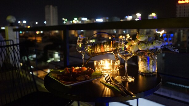 Ein Tisch mit einer Flasche, Gläsern, einer Kerze und einem Blumenstrauß auf einem Balkon mit Blick auf die beleuchtete Stadt bei Nacht.
