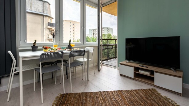 A modern living room with a dining area, a large TV, and a balcony with a view of buildings. - Warsaw Concierge Silver Apartment (Warsaw)