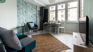 A modern living room with a sofa, a dining table with chairs, a TV, and a large window with a view of buildings. - Warsaw Concierge Silver Apartment (Warsaw)