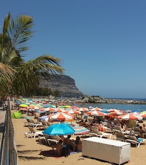 Une plage parsemée de nombreux parasols et de chaises longues, une falaise rocheuse en arrière-plan et un ciel bleu dégagé.