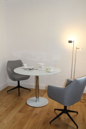 Een ronde glazen tafel met twee grijze stoelen, een vloerlamp en een witte muur.