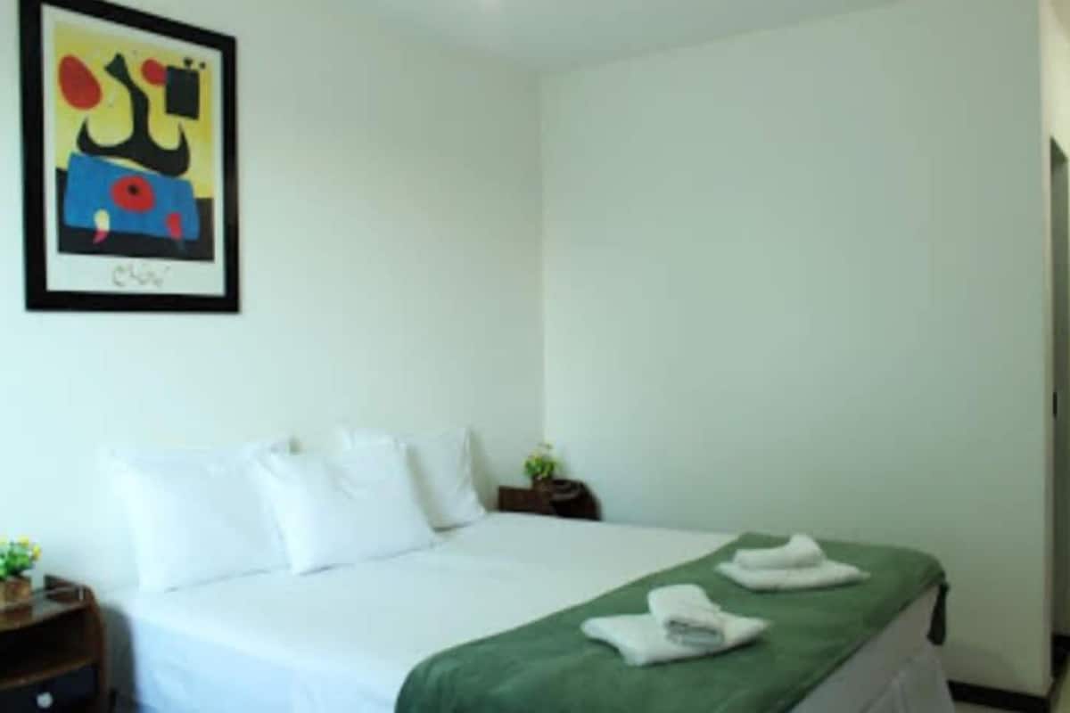 Une chambre d’hôtel avec un lit, des oreillers, une couverture verte, un tableau abstrait encadré et une petite table sur laquelle est posée une plante.