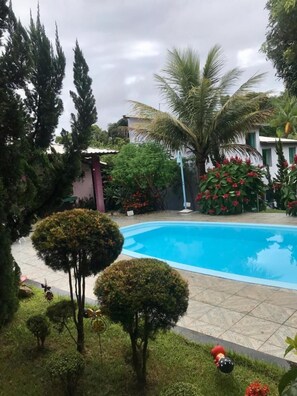 A swimming pool surrounded by well-maintained bushes and trees, with a small building in the background. - Pousada Cantim de BH (Belo Horizonte)