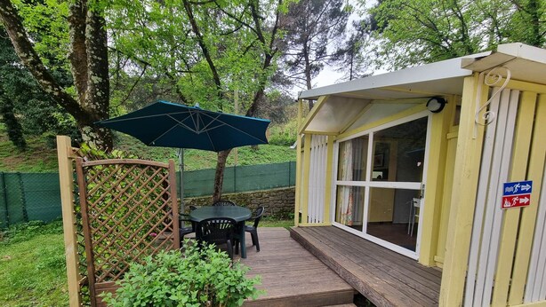 Une terrasse en bois avec une table et des chaises, un espace salon couvert et un petit bâtiment doté d’une fenêtre.