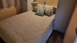 Une petite chambre à coucher accueillante, avec une couvre-lit à motifs, une table de chevet dotée d’une lampe et une petite étagère ornée d’une plante.