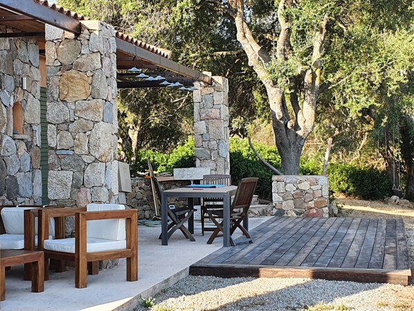 Un patio al aire libre de piedra con mesa y sillas de madera, rodeado de follaje y árboles.