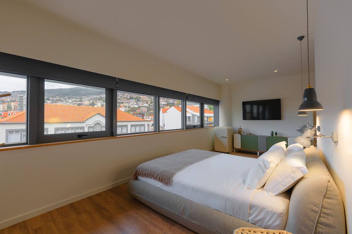 A bedroom with a large bed, a nightstand, a lamp, a TV, and a view of buildings through the window.