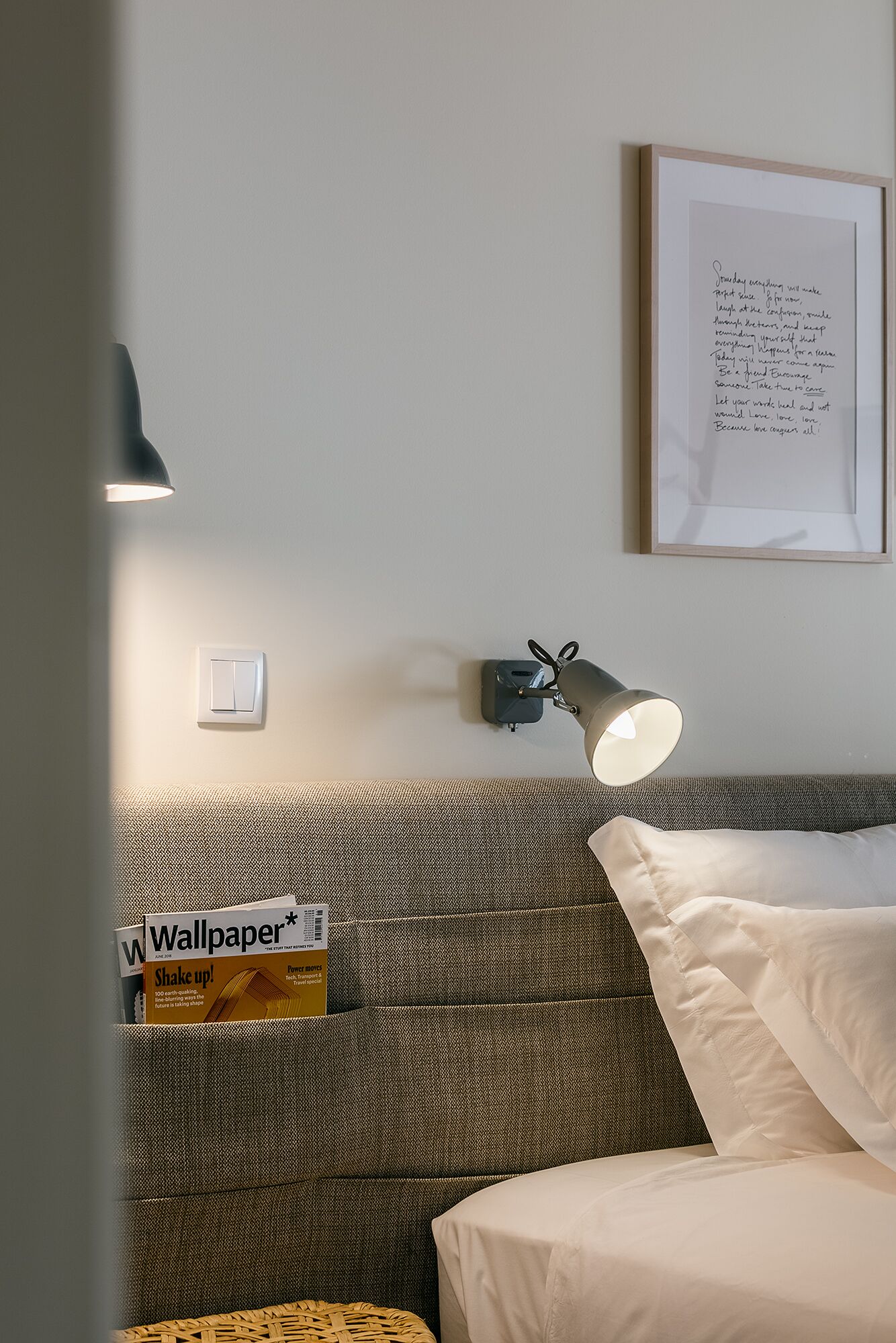 A hotel room with a bed, a wall-mounted lamp, a framed inspirational quote, and a magazine on the headboard.
