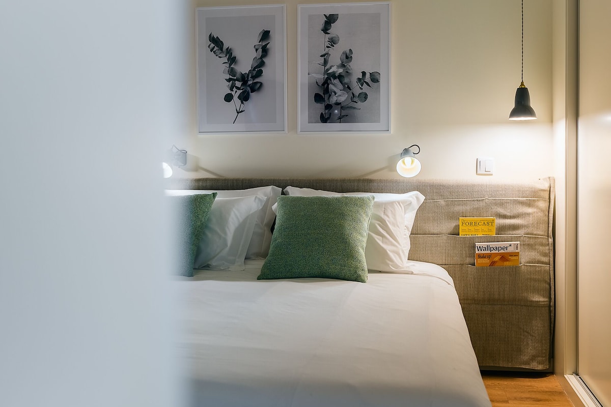 A neatly made bed with white and green pillows, a wall-mounted light, and two framed artworks above it.