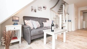 A modern living room with a grey sofa, white coffee table, and a white side table with a lamp. A staircase is visible in the background. - Blinkfüür No.25 (Krummhoern)