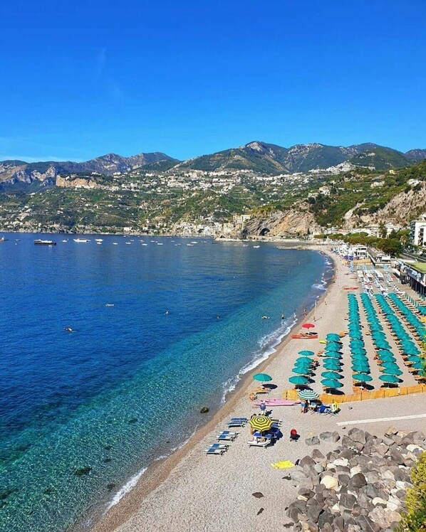 A beach with clear blue water, rows of umbrellas, and a rocky shoreline.