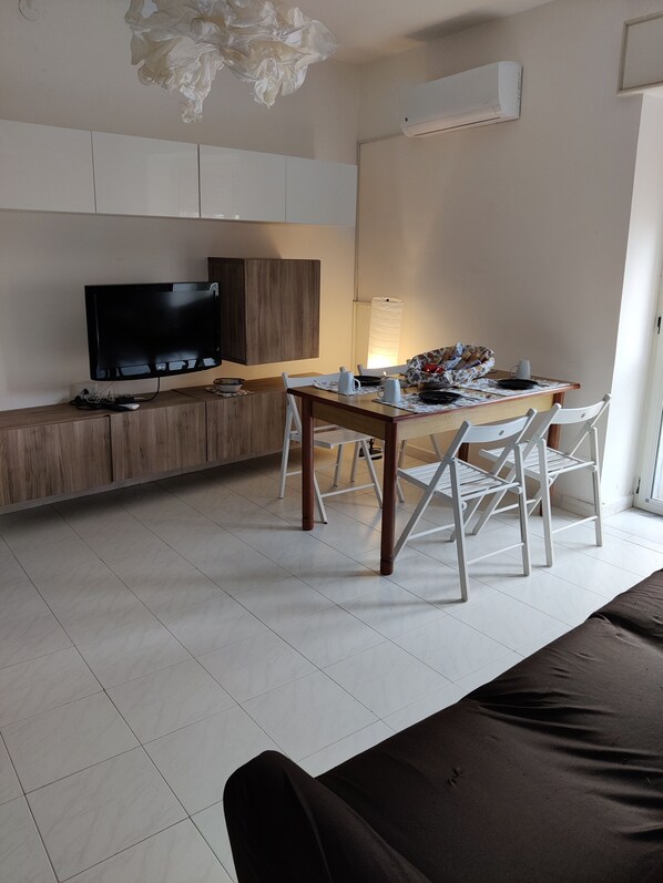 A modern kitchen with a flat-screen TV, wooden cabinets, a dining table with chairs, and a hanging light fixture.