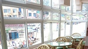 Un restaurante con sillas de mimbre y cojines verdes, amplias ventanas con vistas a la calle y suelo de baldosas.