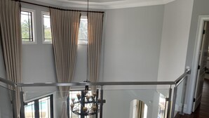 Una habitació àmplia amb un sostre alt, grans finestres amb cortines, un penjol i vistes cap a l'exterior.