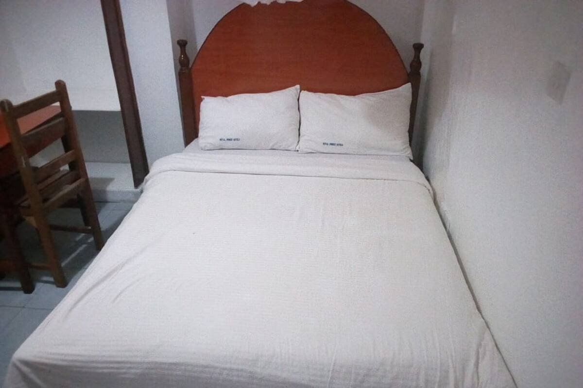 A single bed with a wooden headboard and white bedding in a room with white walls and a wooden chair.
