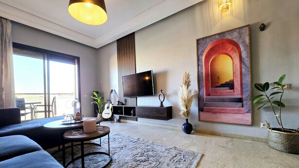 A modern living room with a sofa, coffee table, and TV. There's a large window, a staircase, and a potted plant. - Apartment Bagatelle (Marrakech)