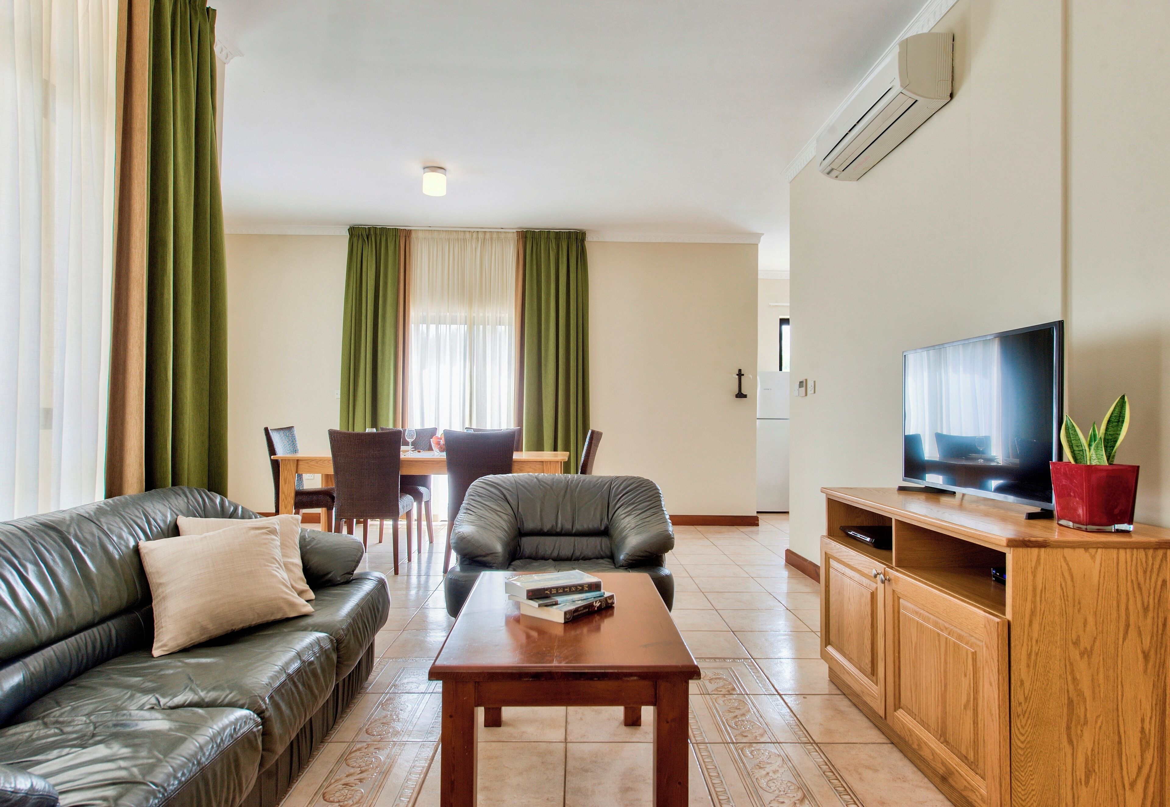 A living room with a leather sofa, a wooden coffee table, a TV on a wooden stand, and a dining area with a table and chairs.
