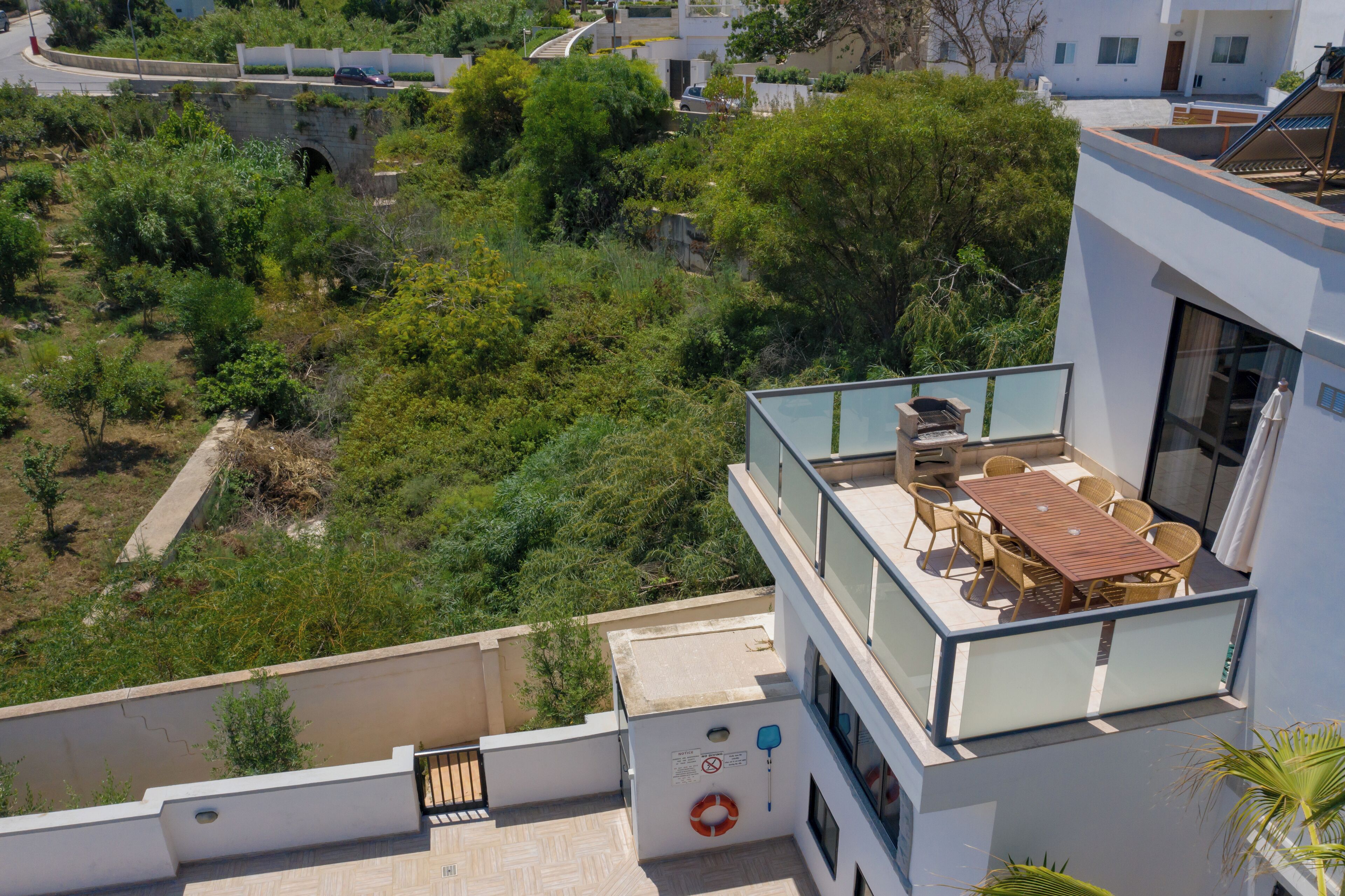 A modern building with a rooftop terrace featuring a table and chairs, surrounded by lush greenery and a stone bridge in the background.
