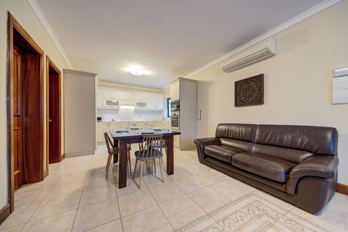 A living room with a brown leather sofa, a dining table with chairs, and a kitchen in the background.