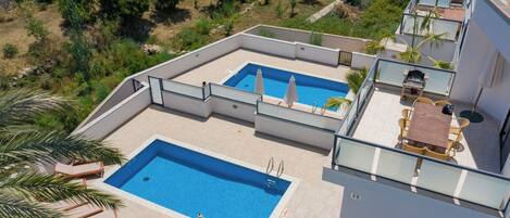 A rooftop pool area with two pools, lounge chairs, and a table with umbrellas.