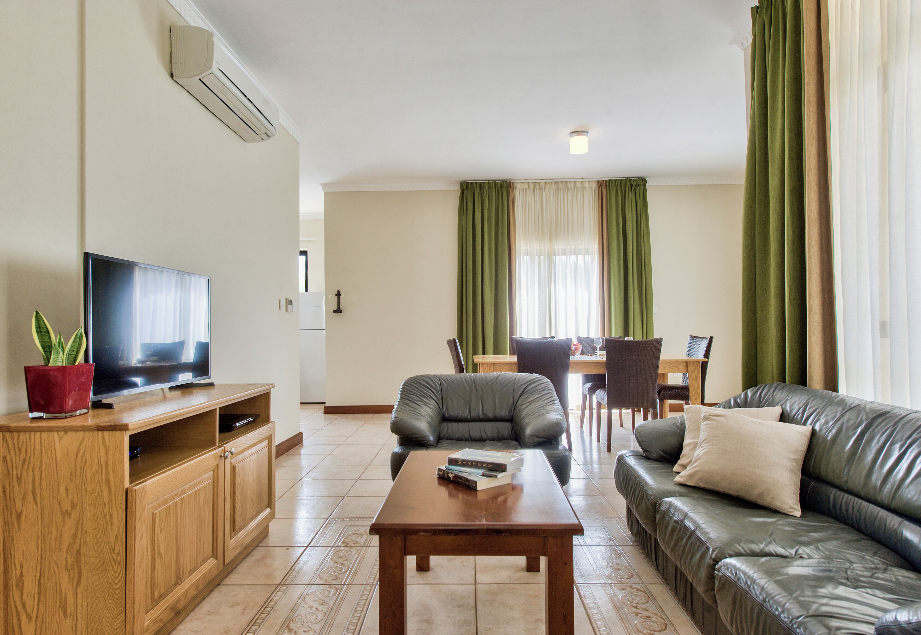 A living room with a sofa, a coffee table, a TV on a wooden stand, a dining table with chairs, and curtains.