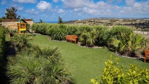 A well-maintained garden with a play area, including a slide and a wooden bench, surrounded by lush greenery and a clear view of the landscape. - Villa Munqar (Zurrieq)