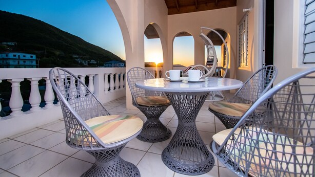 Ein Balkon mit rundem Tisch und vier Stühlen, der einen malerischen Blick auf eine Hügel- und Stadtlandschaft bei Sonnenuntergang bietet.