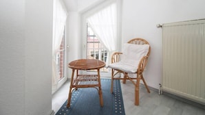 A small balcony with a wicker chair and a round table, white curtains, and a view of a building outside.