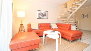 A modern living room with a sofa, ottoman, and a white coffee table. There is a spiral staircase to the right and a framed picture on the wall.