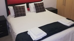 A neatly made bed with a red headboard, two pillows, and a black throw blanket. There are two bedside tables with lamps, a wooden wardrobe, and a wall decoration above the bed. - Luxury, comfortable and secure home away from home for the best JHB experience (Sandton)