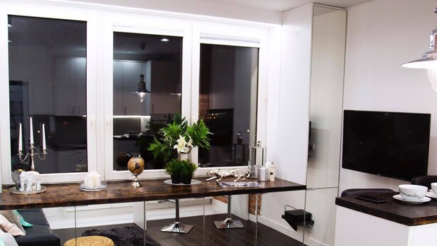 Un salón moderno con suelo de madera oscura, un sofá negro y una alfombra negra. Hay una zona de cocina con ventana, un televisor y una planta en la encimera.