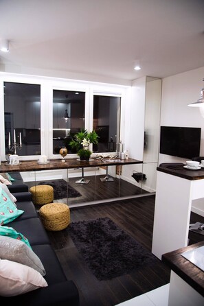 A modern living room with a dark wood floor, black sofa, and a black rug. There is a kitchen area with a window, a TV, and a plant on the counter. - Warsaw Concierge Modern Studio (Warsaw)