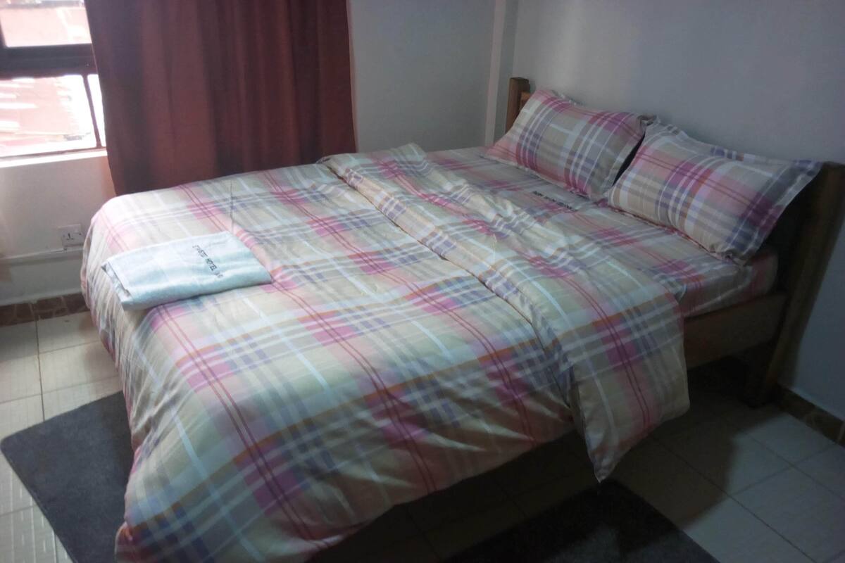 Una cama con colcha y almohadas a cuadros, una alfombra oscura y una ventana con cortinas.