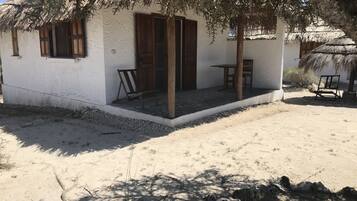 A white building with wooden shutters, a thatched roof, and a small porch with two wooden chairs.