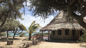 A rustic thatched-roof hut with a stone exterior, surrounded by trees and a sandy area, overlooking a body of water. - Manga Lodge (Andavadoaka)