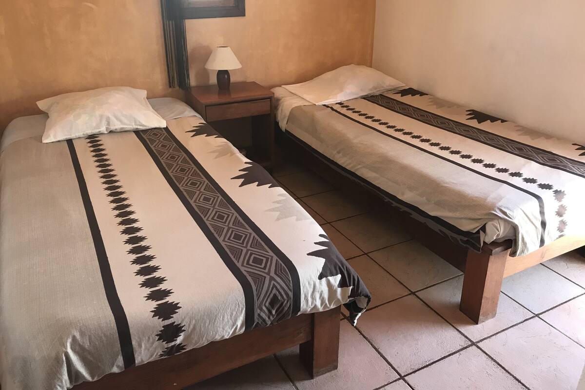 Two single beds with patterned bedding, a nightstand with a lamp, and a window in a room with plain walls.