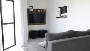 A modern living room with a grey sofa, a flat-screen TV mounted on the wall, and two black speakers. There are framed pictures on the wall, and a door with curtains is visible. - APARTMENT ON THE BEACH (Bessa)