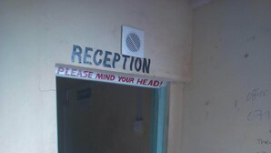 A reception area with a sign that reads "RECEPTION" and "PLEASE MIND YOUR HEAD!" A blue plastic chair with a logo is in the foreground. - Sunset Guest House (Meru)