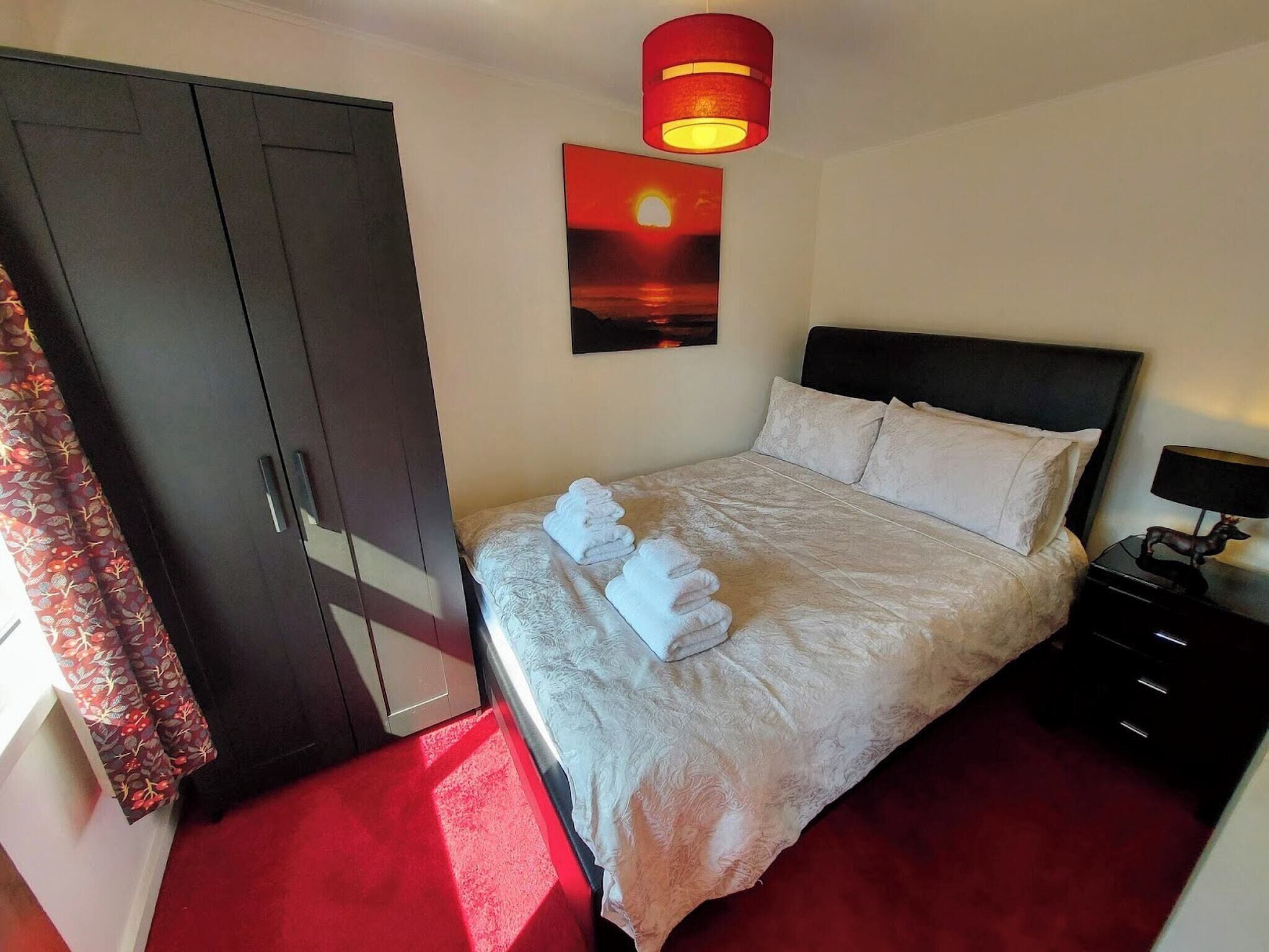 A hotel room with a bed, a nightstand, a lamp, a wardrobe, a painting of a sunset, and a red carpet.
