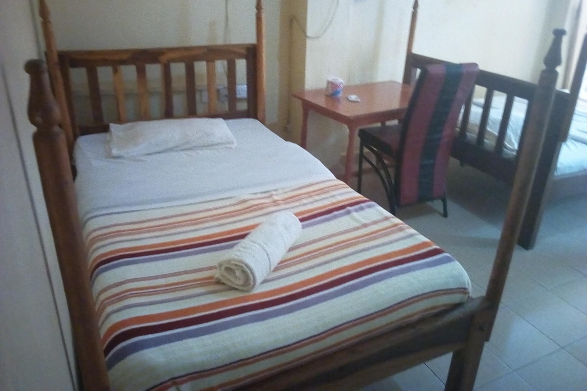 Un lit simple avec une couvre-lit à rayures et une serviette enroulée, une tête de lit en bois, une petite table avec une chaise et une fenêtre avec des rideaux.