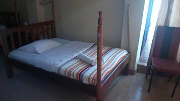 A single bed with a wooden headboard and a striped bedspread. There is a chair to the right and a window with curtains.
