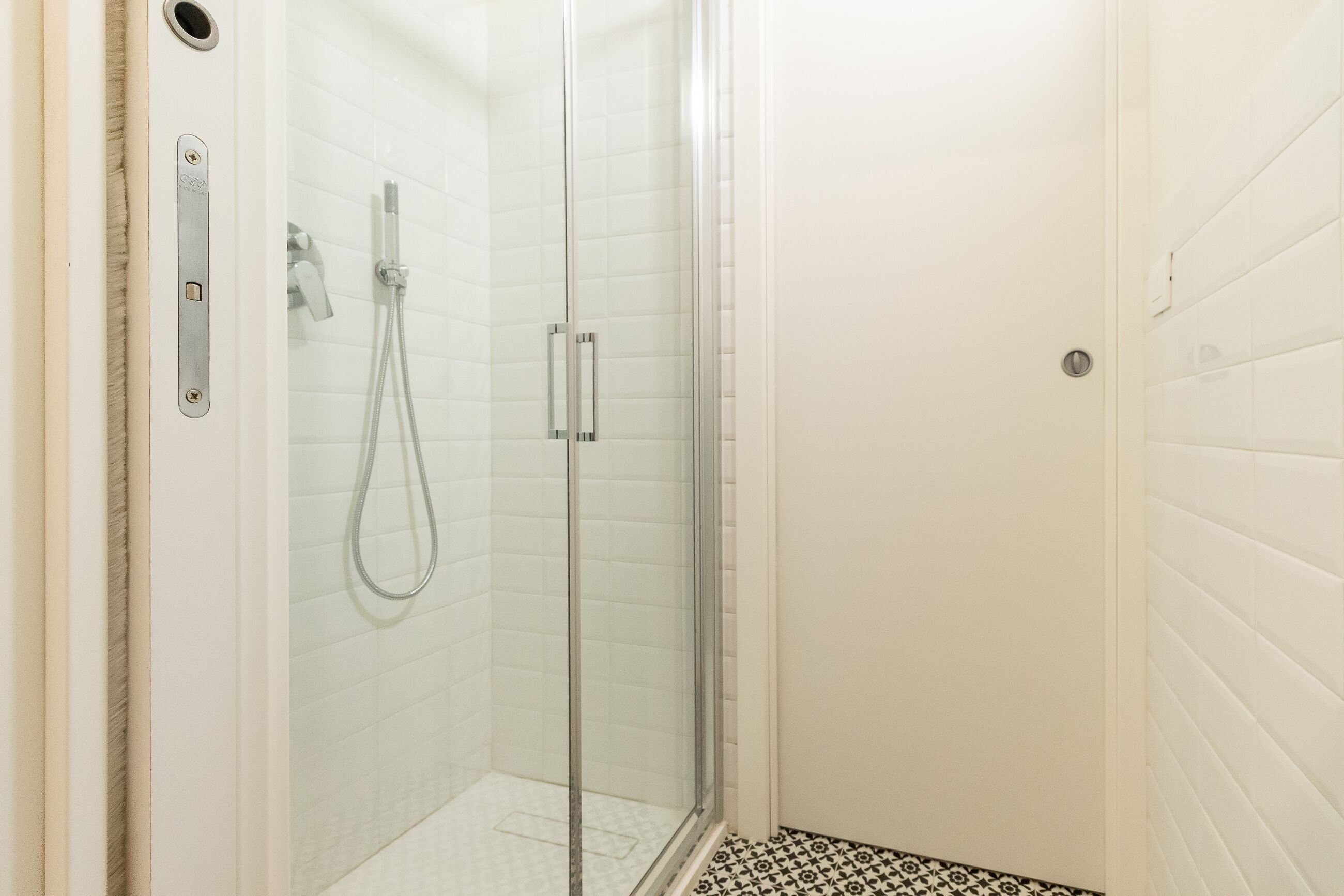 Una ducha con paredes de vidrio, pared con azulejos blancos y suelo con estampado blanco y negro.