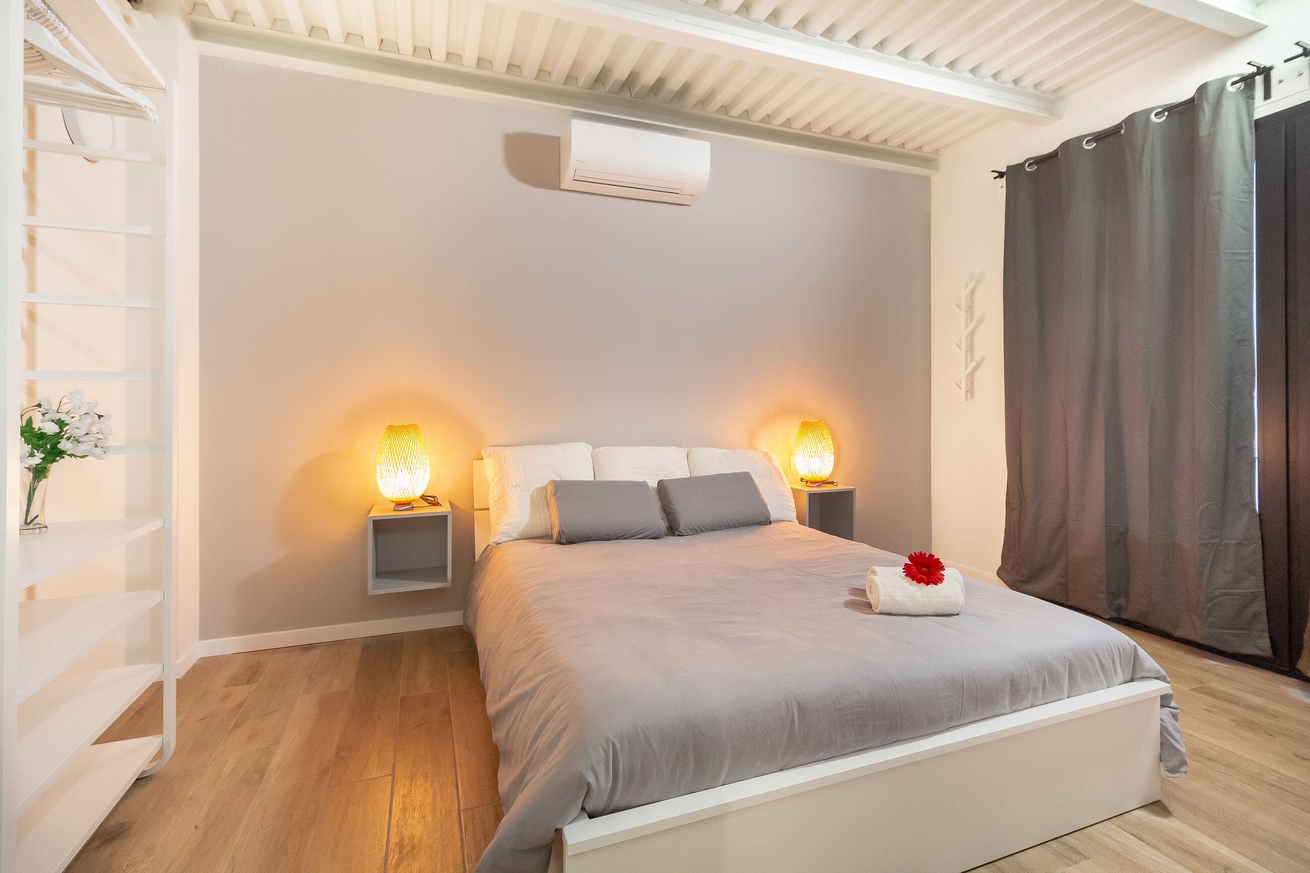 A bedroom with a bed, bedside lamps, a wall-mounted air conditioner, and a window with curtains.