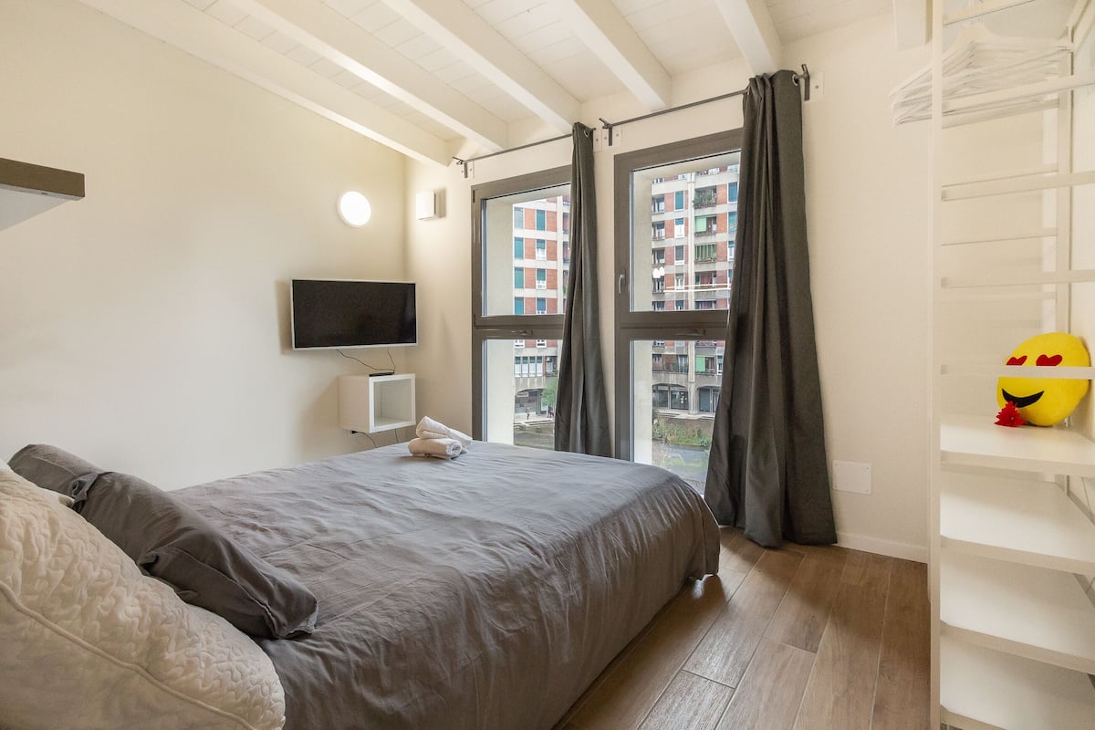 A bedroom with a bed, a TV, a window with curtains, and a view of buildings outside.