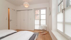 A bedroom with a bed, a rocking chair, a wardrobe, and a window with blinds. - Ferienhaus DeichGalerie89 (Krummhoern)