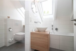 A modern bathroom with a skylight, a toilet, a sink with a vessel basin, and a bathtub. - Ferienhaus DeichGalerie89 (Krummhoern)