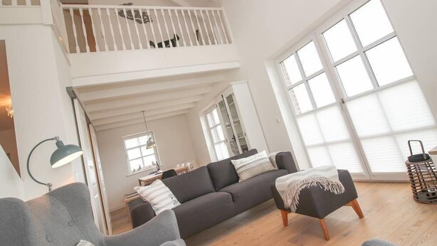 A spacious living area with a high ceiling, a grey sofa, a black ottoman, and a white railing on the upper level. - Ferienhaus DeichGalerie89 (Krummhoern)