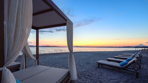 Camera d'hotel affacciata sulla spiaggia, con vista sul tramonto sull'acqua, una zona relax coperta con poltrone e una spiaggia sabbiosa deserta.