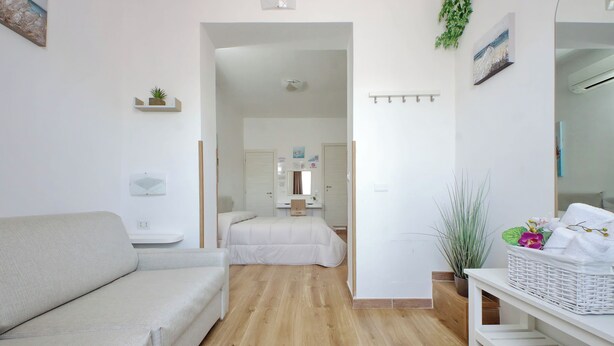 A modern, minimalist room with a wooden floor, a white sofa, a bed, a small table, and a wall-mounted shelf with a plant. - B&B Felicioni (Fiumicino)